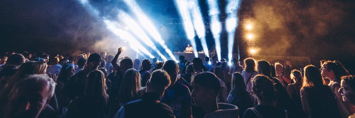 Rhein in Flammen Lahnstein Menschen stehen dicht zusammen vor einer Open-Air-Bühne und feiern ein Konzert bei Nacht. Helle Scheinwerferstrahlen und Nebel durchbrechen den dunklen Himmel, während ein DJ oder Künstler auf der Bühne auftritt und die Lichtshow die Festivalatmosphäre prägt.