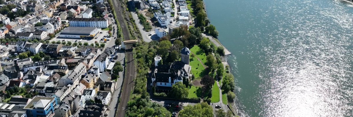 Luftaufnahme von Oberlahnstein am Rhein: Entlang des Flussufers erstrecken sich Wohngebiete, Verkehrswege und Grünanlagen. Im Vordergrund ist das Martinsschloss mit seinem markanten Turm zu sehen, dahinter verläuft die Bahnstrecke parallel zum Rhein. Die Aufnahme zeigt das Stadtgebiet eingebettet zwischen Fluss und bewaldeten Höhenzügen des Mittelrheintals.