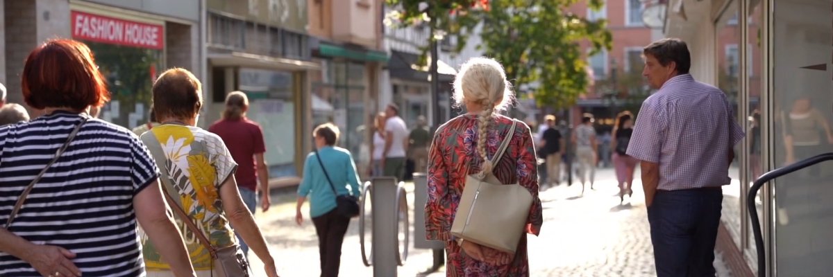 Menschen schlendern an einem sonnigen Tag durch eine Einkaufsstraße in der Lahnsteiner Innenstadt. Zwischen Geschäften und Schaufenstern sind Passantinnen und Passanten unterwegs, während Blumenampeln und das warme Licht eine lebendige und einladende Atmosphäre schaffen. 