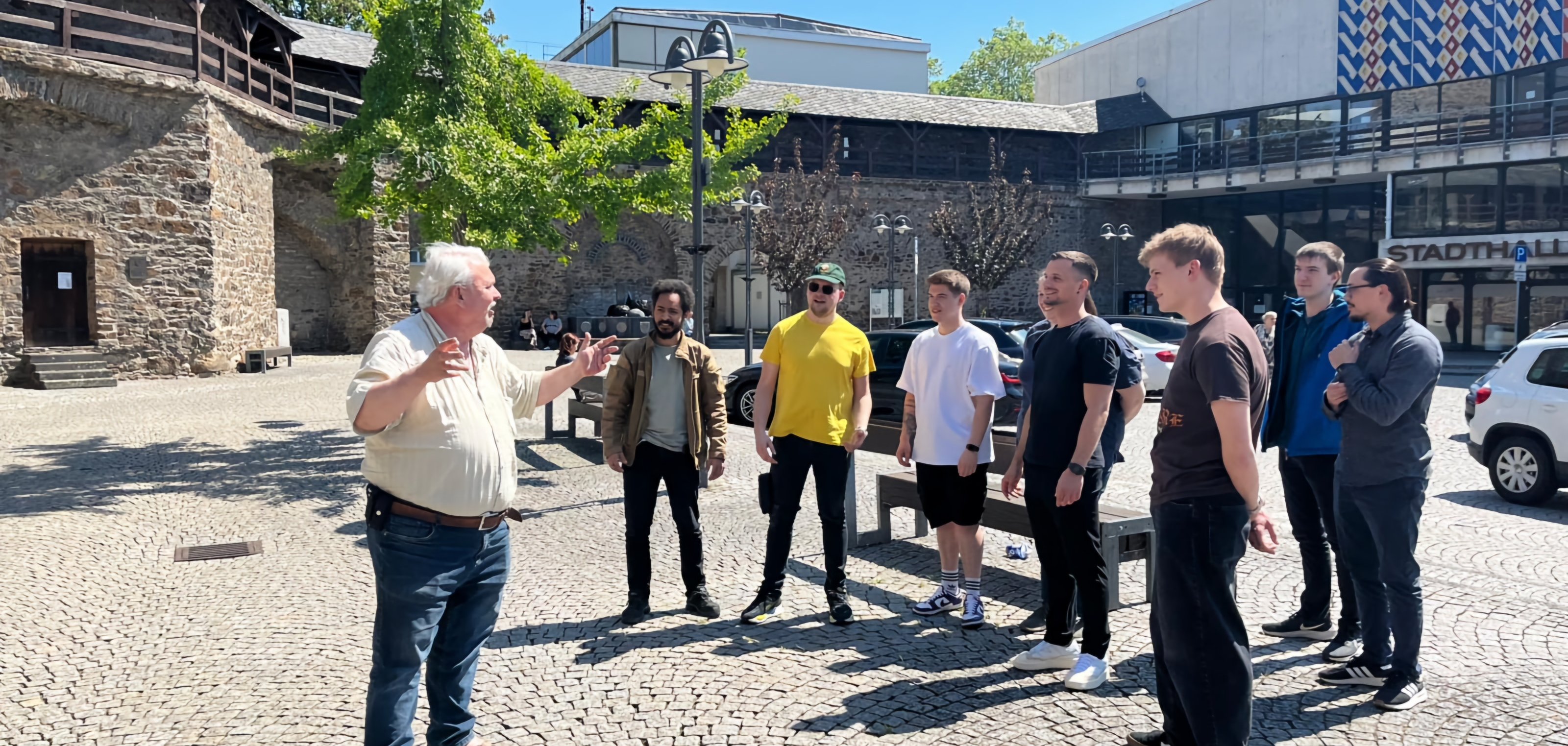 Eine Gruppe junger Erwachsener steht auf dem Salhofplatz in Lahnstein und hört einem Stadtführer zu, der mit ausladenden Gesten die Geschichte des Ortes erläutert. Im Hintergrund sind die Stadtmauer, historische Gebäude und die Stadthalle zu sehen. Die Aufnahme entstand bei sonnigem Wetter während einer Stadtführung durch die Innenstadt.