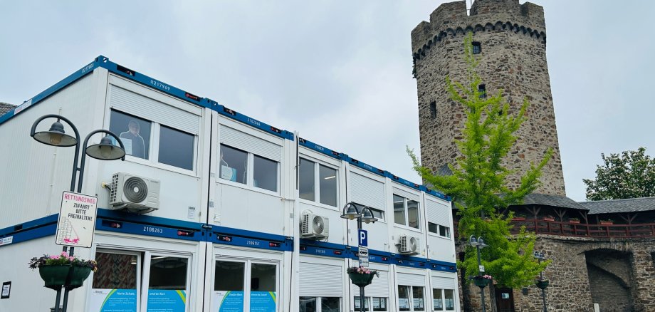 Servicecenter Lahnstein Zweigeschossiger Bürocontainer mit Klimageräten und großen Fenstern steht auf einem gepflasterten Platz neben einem historischen, runden Steinturm mit Zinnen und gehisster Flagge. Umgeben ist der Container von Straßenlaternen und einem jungen Baum.