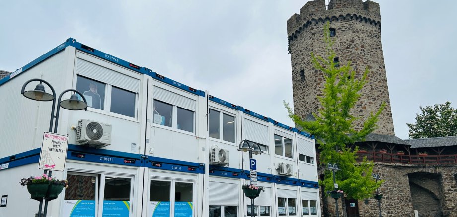 Servicecenter Lahnstein Zweigeschossiger Bürocontainer mit Klimageräten und großen Fenstern steht auf einem gepflasterten Platz neben einem historischen, runden Steinturm mit Zinnen und gehisster Flagge. Umgeben ist der Container von Straßenlaternen und einem jungen Baum.
