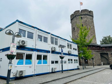 Servicecenter Lahnstein Zweigeschossiger Bürocontainer mit Klimageräten und großen Fenstern steht auf einem gepflasterten Platz neben einem historischen, runden Steinturm mit Zinnen und gehisster Flagge. Umgeben ist der Container von Straßenlaternen und einem jungen Baum.
