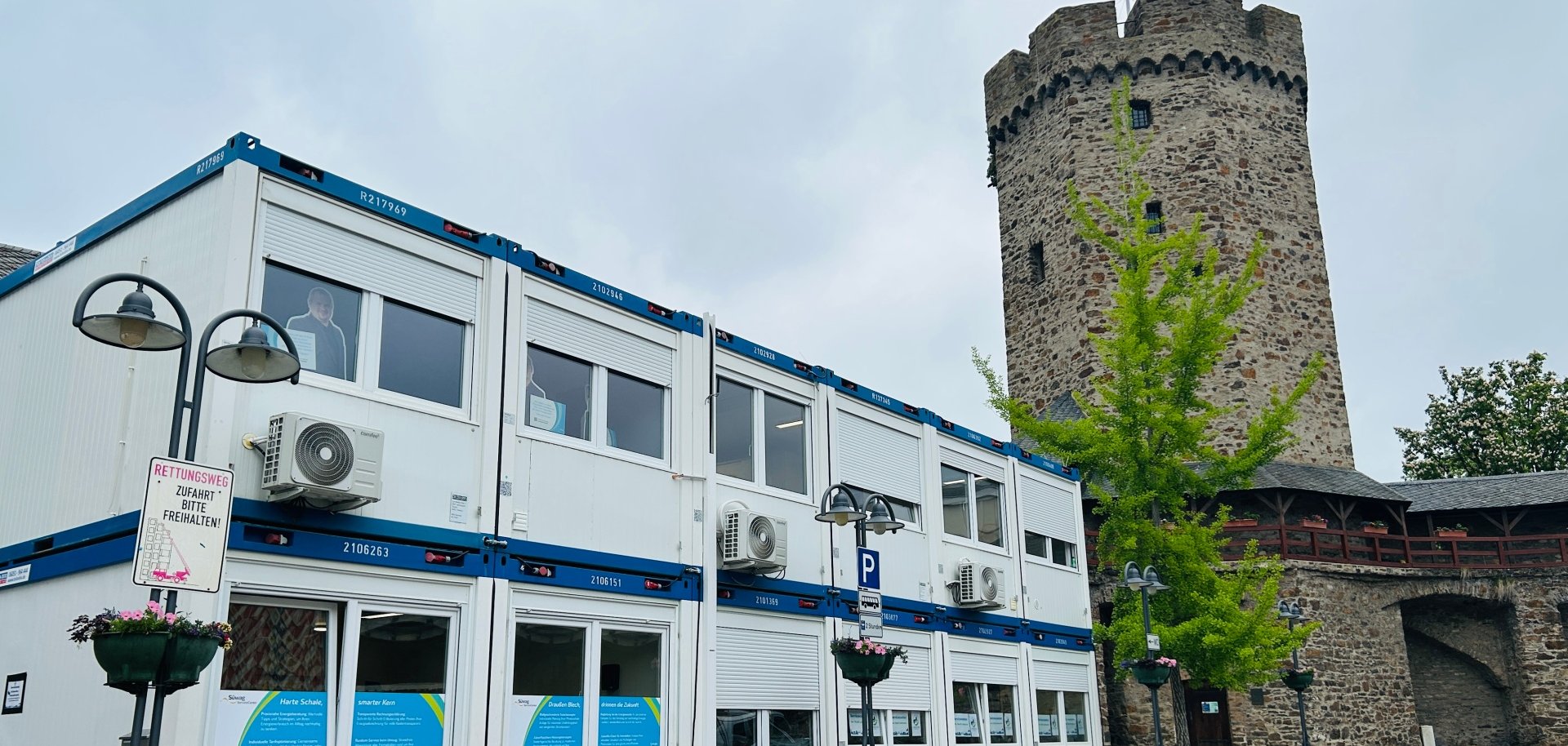 Zweigeschossiger Bürocontainer mit Klimageräten und großen Fenstern steht auf einem gepflasterten Platz neben einem historischen, runden Steinturm mit Zinnen und gehisster Flagge. Umgeben ist der Container von Straßenlaternen und einem jungen Baum.