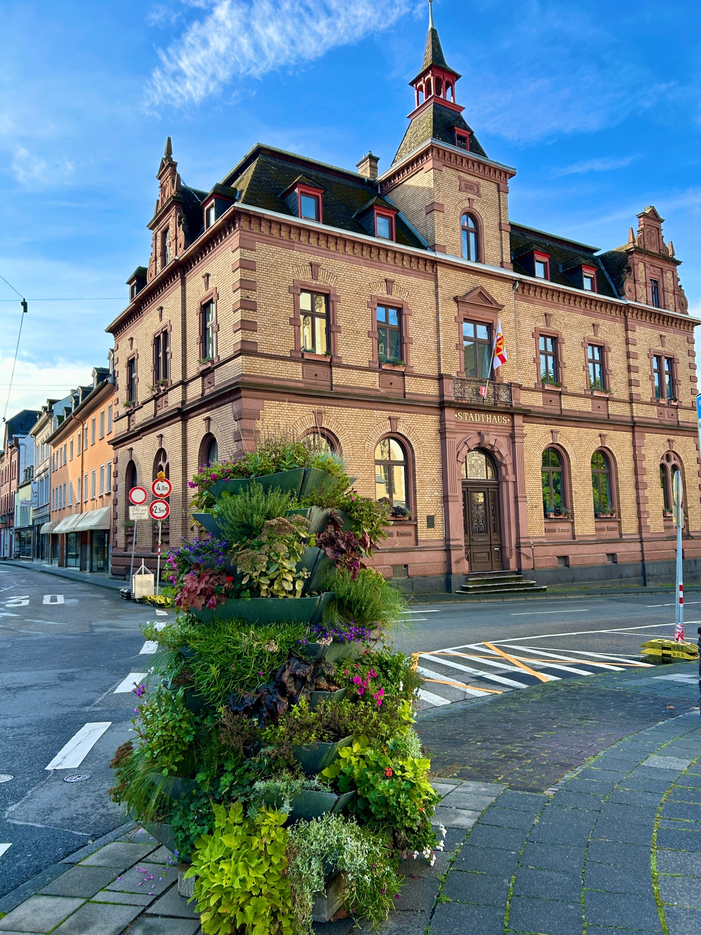 Historisches Rathausgebäude aus hellem Backstein mit dekorativen roten Sandsteinelementen und einem spitzen Turm unter blauem Himmel; davor steht ein bepflanztes, mehrstöckiges Blumenarrangement an einer ruhigen Straßenecke.