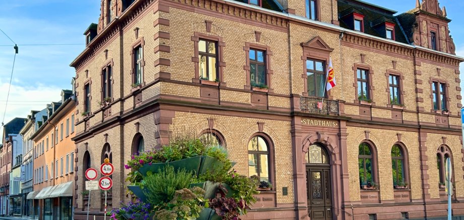 Historisches Rathausgebäude aus hellem Backstein mit dekorativen roten Sandsteinelementen und einem spitzen Turm unter blauem Himmel; davor steht ein bepflanztes, mehrstöckiges Blumenarrangement an einer ruhigen Straßenecke.
