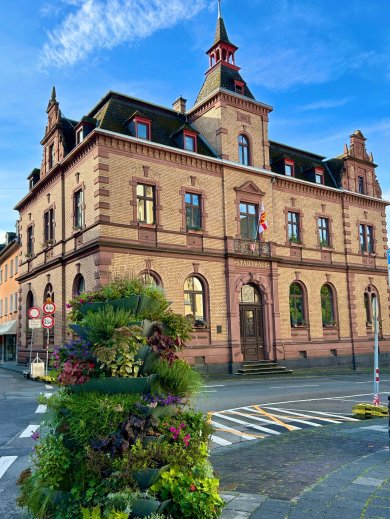 Rathaus Kirchstraße Historisches Rathausgebäude aus hellem Backstein mit dekorativen roten Sandsteinelementen und einem spitzen Turm unter blauem Himmel; davor steht ein bepflanztes, mehrstöckiges Blumenarrangement an einer ruhigen Straßenecke.