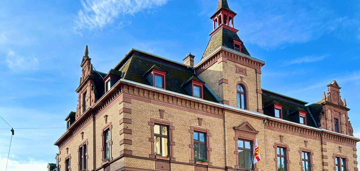 Historisches Rathausgebäude aus hellem Backstein mit dekorativen roten Sandsteinelementen und einem spitzen Turm unter blauem Himmel; davor steht ein bepflanztes, mehrstöckiges Blumenarrangement an einer ruhigen Straßenecke.